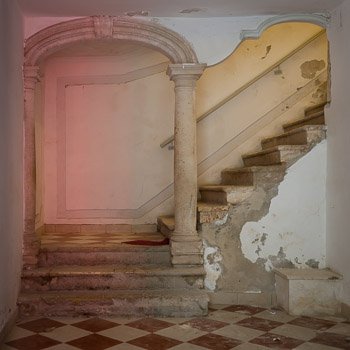 In Its Best Light - Interior Architectural Photography, Dubrovnik, Croatia, by Nat Coalson. The foyer of an old building in Dubrovnik, Croatia is lit with bright colors, temporarily brightening the appearance of the peeling paint and crumbling walls of a staircase leading upward to the unseen floors above.